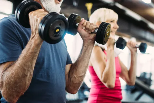 Two older adults lift weights inside of a gym.